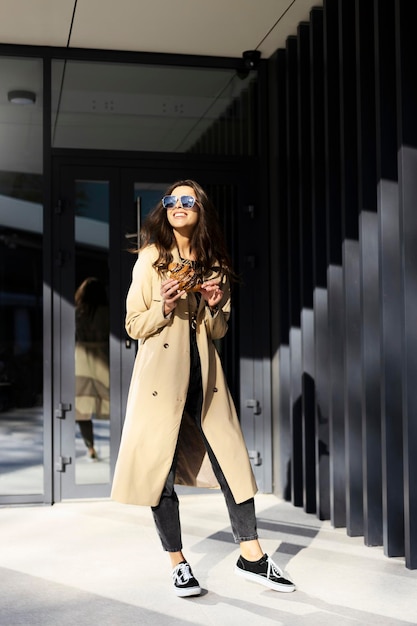 Cheerful smiling brunette in sunglasses with a croissant in her hands is standing near a modern building Beautiful girl in a casual style