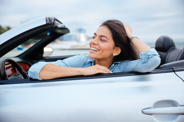 Cheerful smiling brunette woman resting in her cabriolet parked on the beach