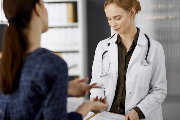 Cheerful smiling female doctor and patient woman discussing current health examination while sitting in clinic. Perfect medical service in hospital. Medicine concept.