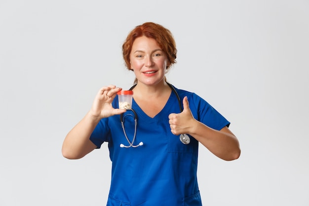 Cheerful smiling female meical worker, doctor in scrubs showing container with vitamins or medication, recommend pills, showing thumbs-up in approval.