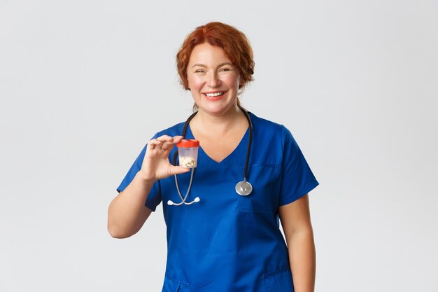 Cheerful smiling female meical worker, doctor in scrubs showing container with vitamins or medication, recommend pills, standing grey background.