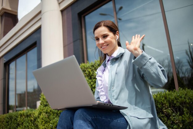 Cheerful smiling lady waves into the laptop screen in the city Video call conference lifestyle