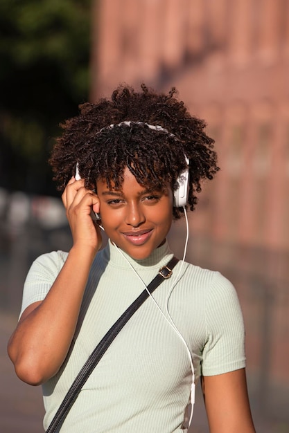 Cheerful smiling Latin American woman with curly hair listening to music on smartphone wears earphones in the street looking at camera