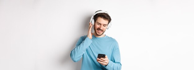 Cheerful smiling man listening music and looking at smartphone reading message on phone standing on