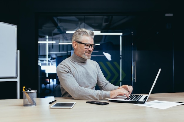 Cheerful and smiling mature businessman working inside office senior gray haired man using laptop at