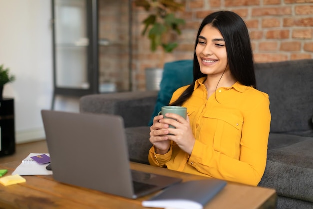 Cheerful spanish lady watching videos on laptop and drinking coffee at home looking at computer