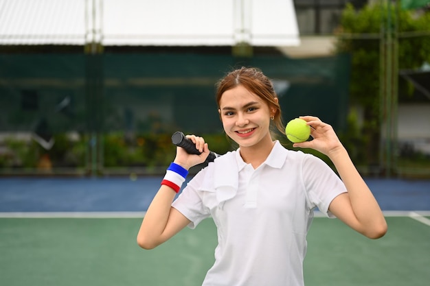 Cheerful sportswoman holding racket and ball standing on the tennis court Fitness sport and competition concept