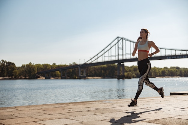 Cheerful sportswoman in tracksuit and sneakers runs along city embankment