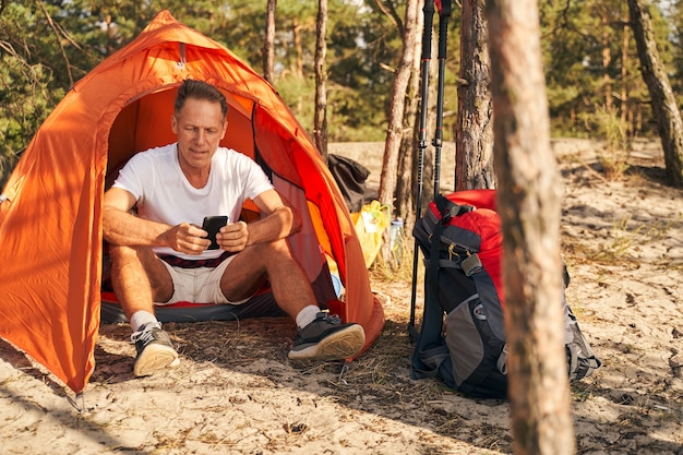 Cheerful sporty man is relaxing in tent and messaging on smartphone after nordic walking in forest