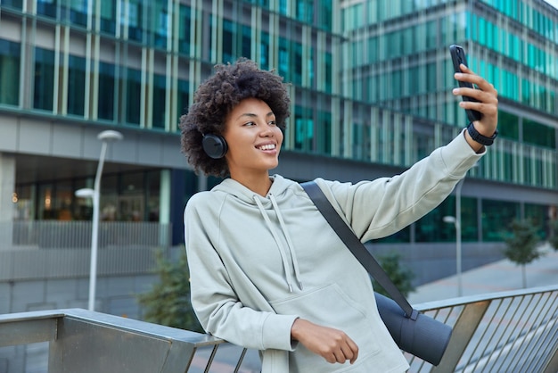 Cheerful sporty woman in hoodie carries yoga mat poses for making selfie with smartphone listens music in wireless headphones stands against modern glass buildings. People wellness sport concept