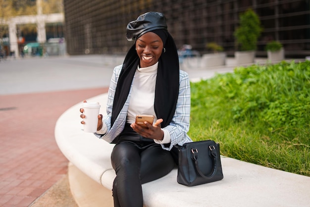 Cheerful stylish black woman in headscarf using smartphone in city