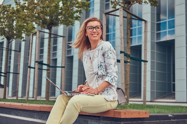 Cheerful stylish fashion blogger relaxing outdoor, working on the laptop, sitting on a bench against a skyscraper.