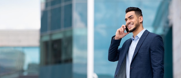 Cheerful successful arab millennial businessman calling by smartphone near building office outdoor