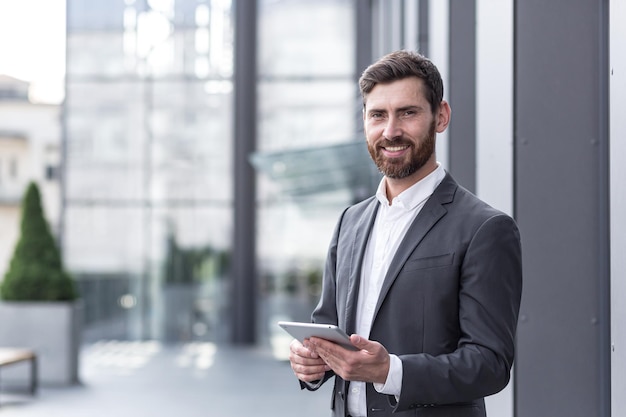 Cheerful and successful businessman works outdoors near the office holds a tablet, smiles reads good news