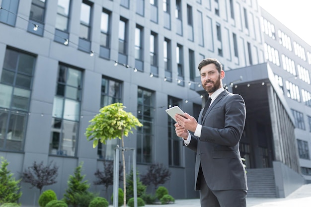 Cheerful and successful businessman works outdoors near the office holds a tablet, smiles reads good news