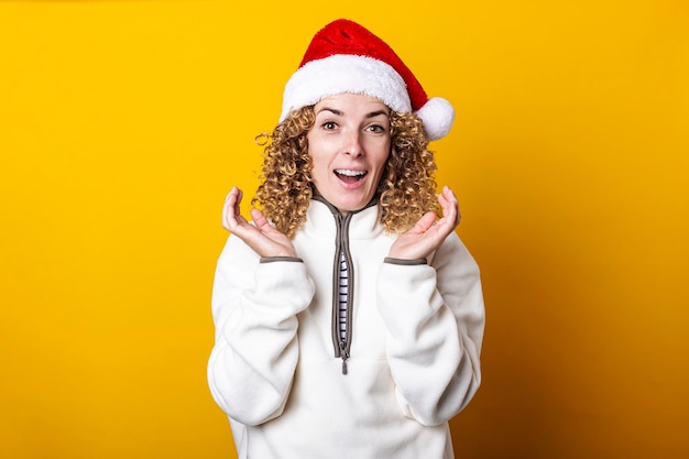 Cheerful surprised curly young woman in santa hat on a yellow background.