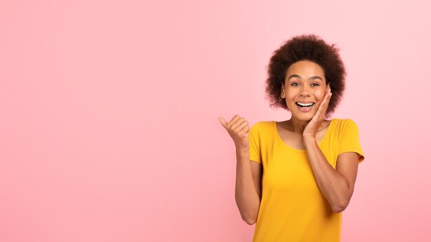 Cheerful surprised millennial african american curly woman in yellow tshirt show finger at empty space