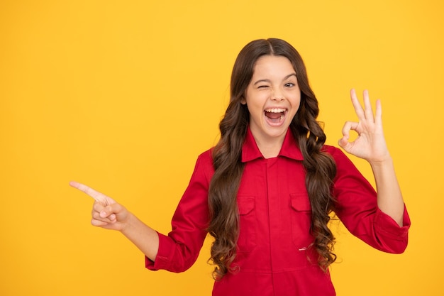 Cheerful teen girl with ok gesture kid presenting product on yellow background child express happiness positive emotions smiling kid pointing finger on copy space happy childhood