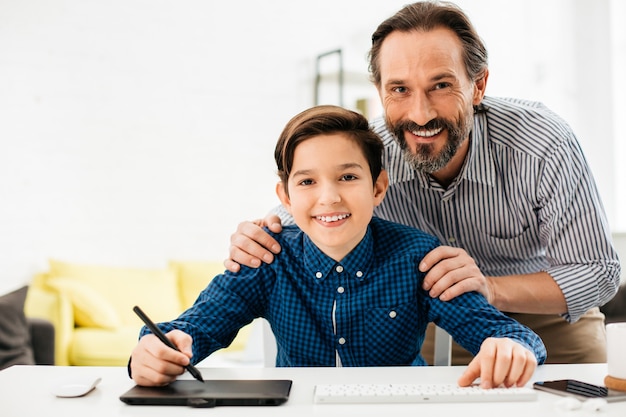 Cheerful teenage boy sitting at the table with stylus in one hand and smiling while positive father standing behind his back