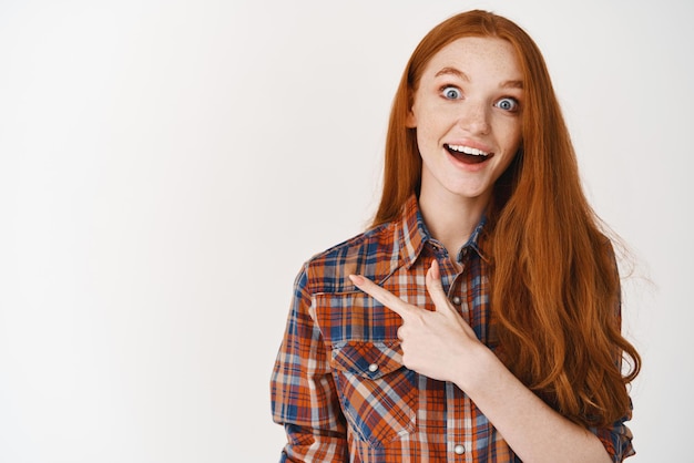 Cheerful teenage girl with red long hair and pale skin smiling and showing logo pointing finger left at promo offer standing over white background