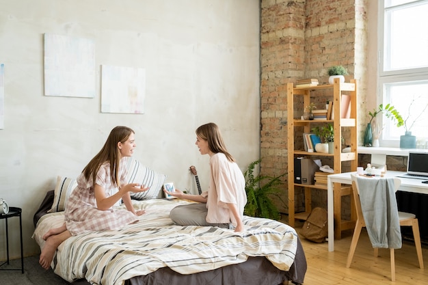 Cheerful teenage twin girls in casualwear discussing funny stuff and laughing while resting on bed in large bedroom