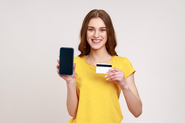 Cheerful teenager girl in yellow casual style tshirt holding and showing empty display smartphone and credit card satisfied with easy mobile banking Indoor studio shot isolated on gray background