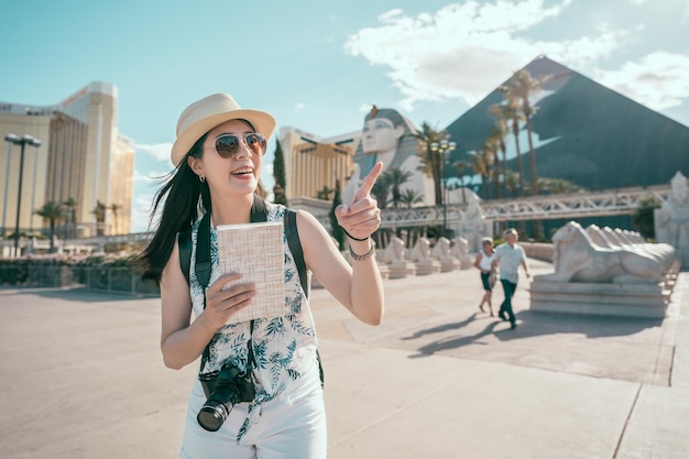 Cheerful tourist with camera standing in front of the sphinx in egypt. young girl traveler enjoying the beauty of the sky and pointing at it. stone statues standing in front of pyramids on sunny day.