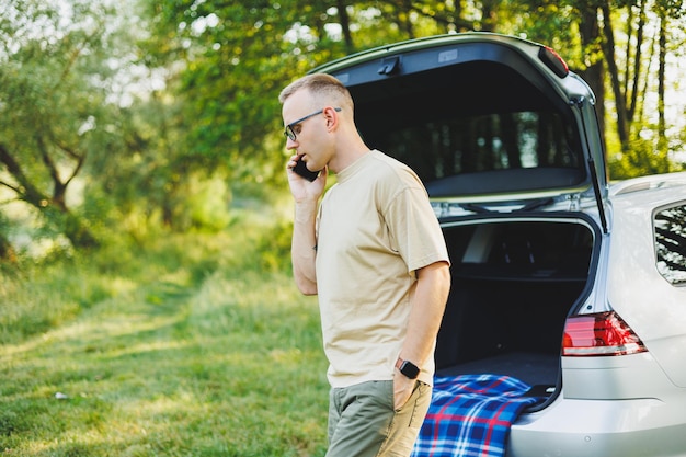 Cheerful traveler young man sitting in open trunk of car working with laptop outdoors concept of business travel vacation