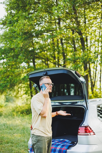 Cheerful traveler young man sitting in open trunk of car working with laptop outdoors concept of business travel vacation