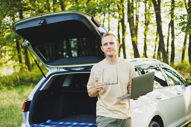 Cheerful traveler young man sitting in open trunk of car working with laptop outdoors concept of business travel vacation