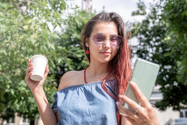 Cheerful trendy woman with red hair drinking coffee at park taking selfie