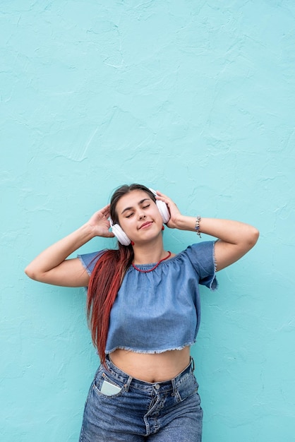 Cheerful trendy woman with red hair listening to the music on blue wall background at street