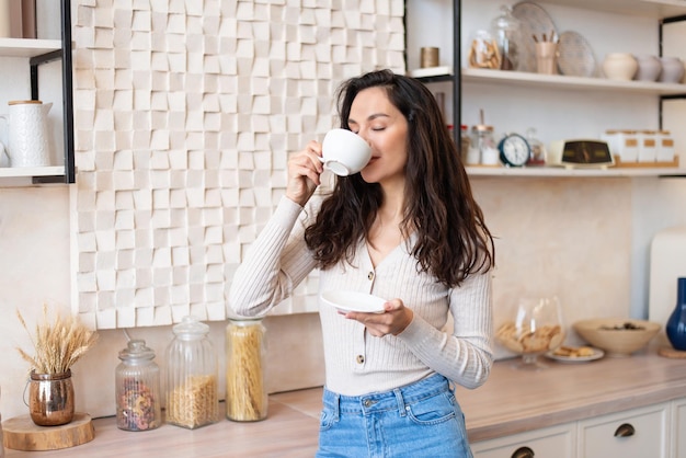 Cheerful woman in casual clothes drinking her favorite hot drink in white cup standing in modern