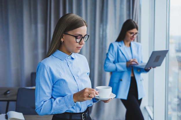 Cheerful woman in classic glasses smiling in free time in office with coffee positive european woman in blue shirt desk with laptop remote work A colleague is in the background selective focus