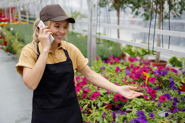 Cheerful woman in florist uniform having a conversation with a client on a smartphone