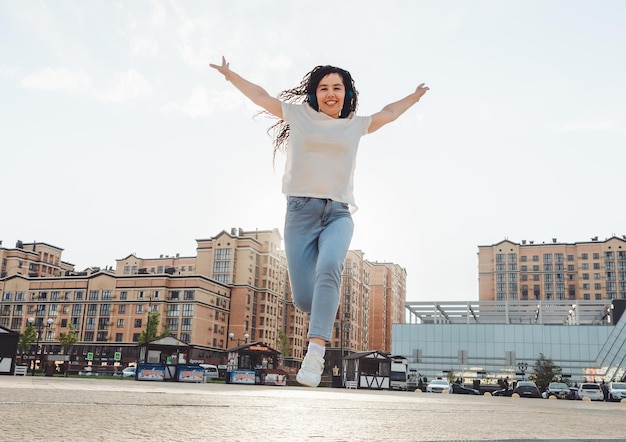 A cheerful woman listens to music through headphones outside on a sunny day a girl with headphones jumps on the street generation z