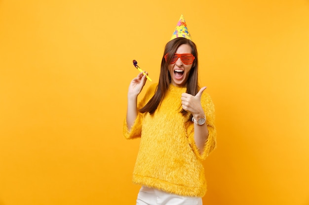 Photo cheerful woman in orange funny glasses, birthday hat with playing pipe showing thumb up, celebrating isolated on bright yellow background. people sincere emotions lifestyle concept. advertising area.