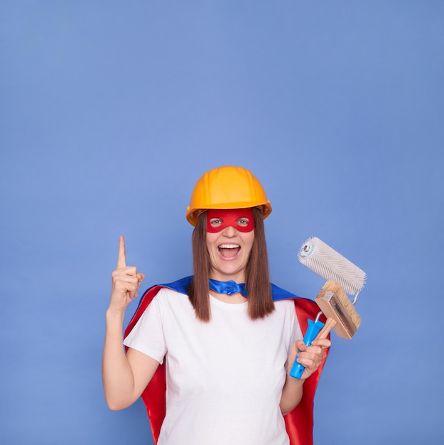 Cheerful woman painter wearing superhero costume and protective helmet holding painting roller and brush isolated over blue background indicating upwards at copy space for advertisement