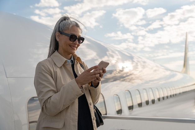 Cheerful woman passenger using phone standing outdoors at airport near plane