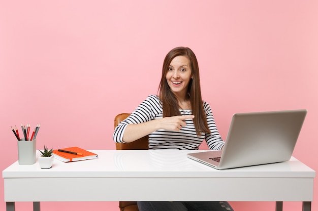 Cheerful woman pointing index finger on contemporary pc laptop while work, sit at desk at office isolated on pastel pink background. Achievement business career concept. Copy space for advertisement.