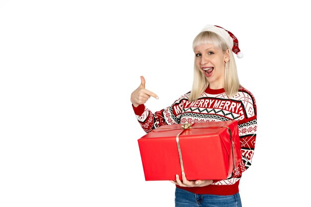 Cheerful woman promoting buying a gift by pointing at it dressed in Christmas sweater and wearing Santa Claus hat Studio background white