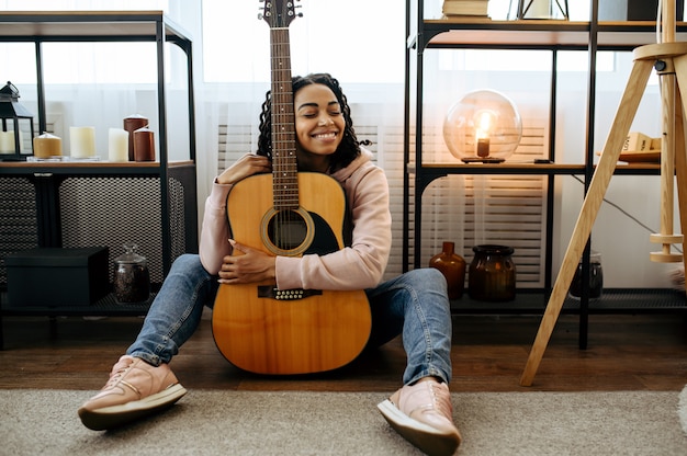 Cheerful woman sitting on the floor and hugs the guitar at home. Pretty lady with musical instrument relax in the room, female music lover resting