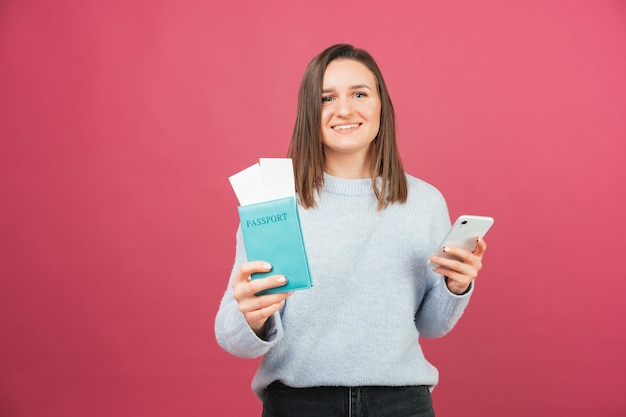 Cheerful woman in sweater holding phone and passport over pink background