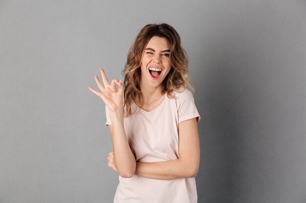 Cheerful woman in t-shirt showing ok sign while with open mouth over grey