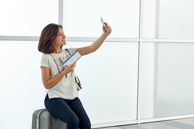 Cheerful woman taking selfie with ticket in airport before departure on white window