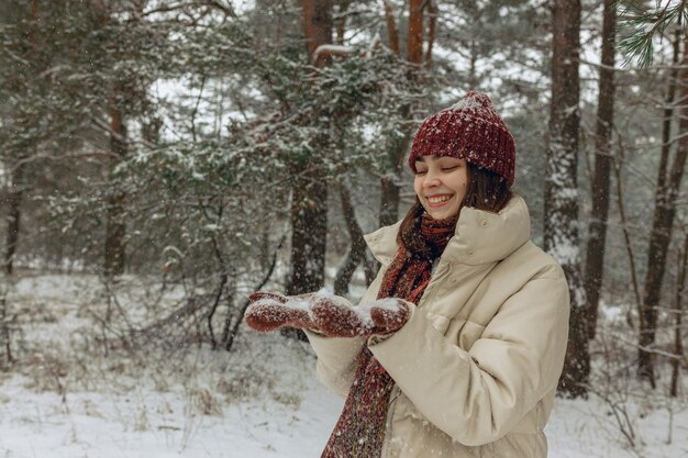 cheerful woman in warm clothes and mittens enjoying winter in snowy forest during snowfall