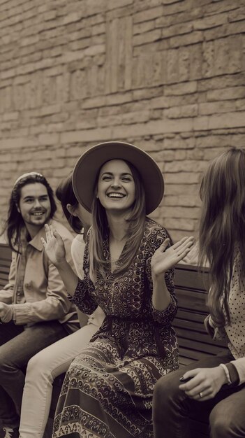 Photo cheerful woman wearing hat sitting amidst friends on bench in front of wall