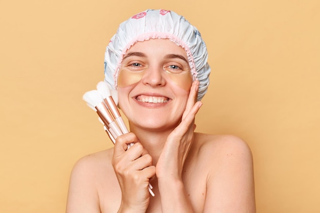 Cheerful woman wearing shower cap standing isolated over beige background posing with under eyes patches holding powder brushes smiling to camera