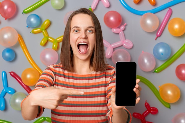 Cheerful woman with brown hair wearing striped dress holding mobile phone with empty screen copy space for advertisement standing against gray wall decorated with colorful balloons