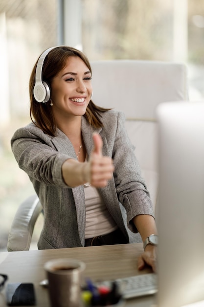 A cheerful woman with headphones showing thumb up during a video call in a modern office.